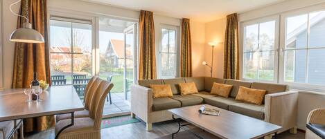 A modern living room with a sofa, coffee table, and chairs. There is a dining table with a lamp and a bottle. Large windows provide a view of the outside.