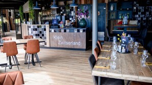 Ein Restaurantinneres mit Holzboden, braunen Ledersesseln und einem langen Esstisch, gedeckt mit Gläsern und Besteck. Ein Schild mit der Aufschrift 'Klein Schweizland' ist sichtbar.