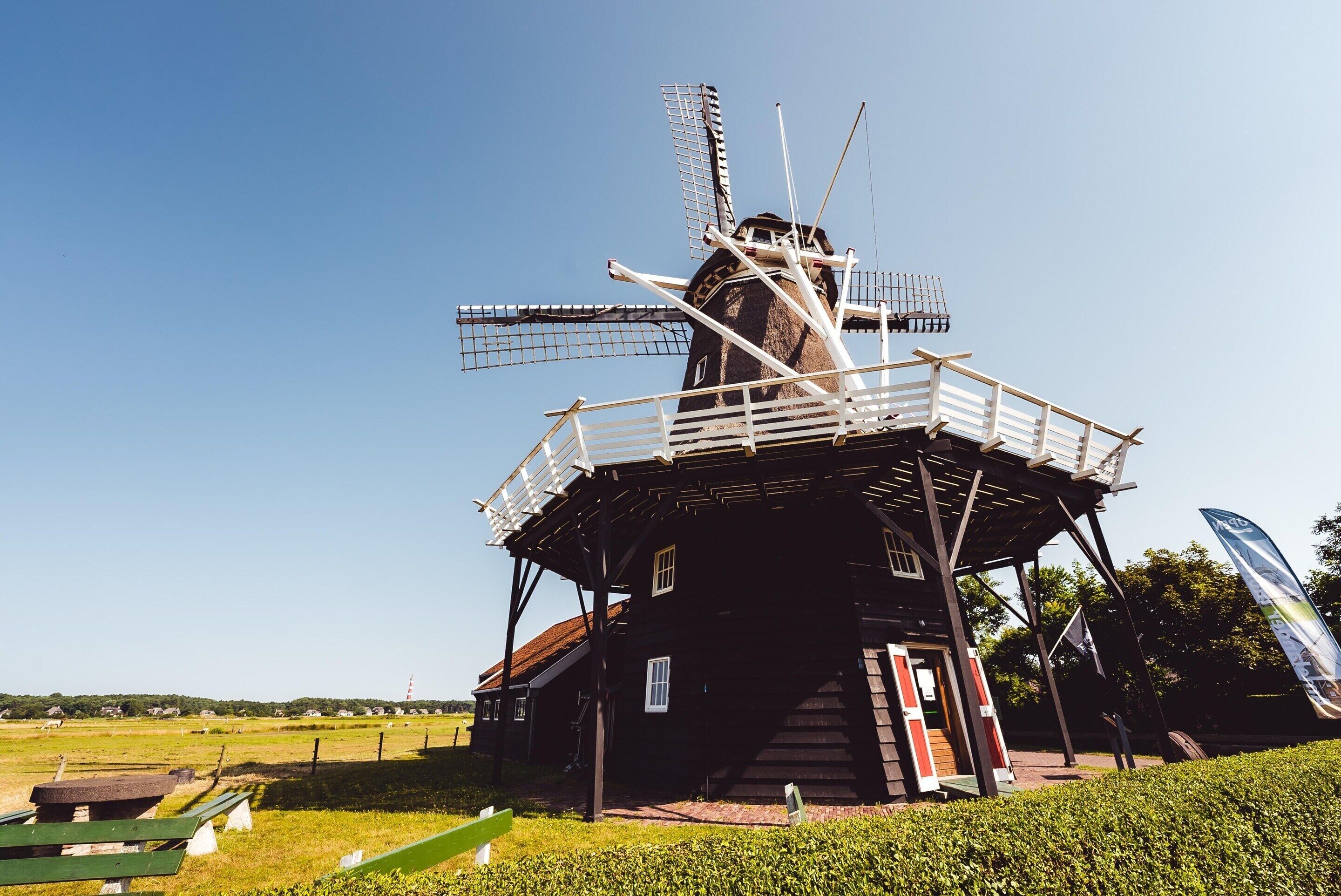 A traditional windmill with a wooden structure and a metal roof, surrounded by a grassy area and a clear sky.