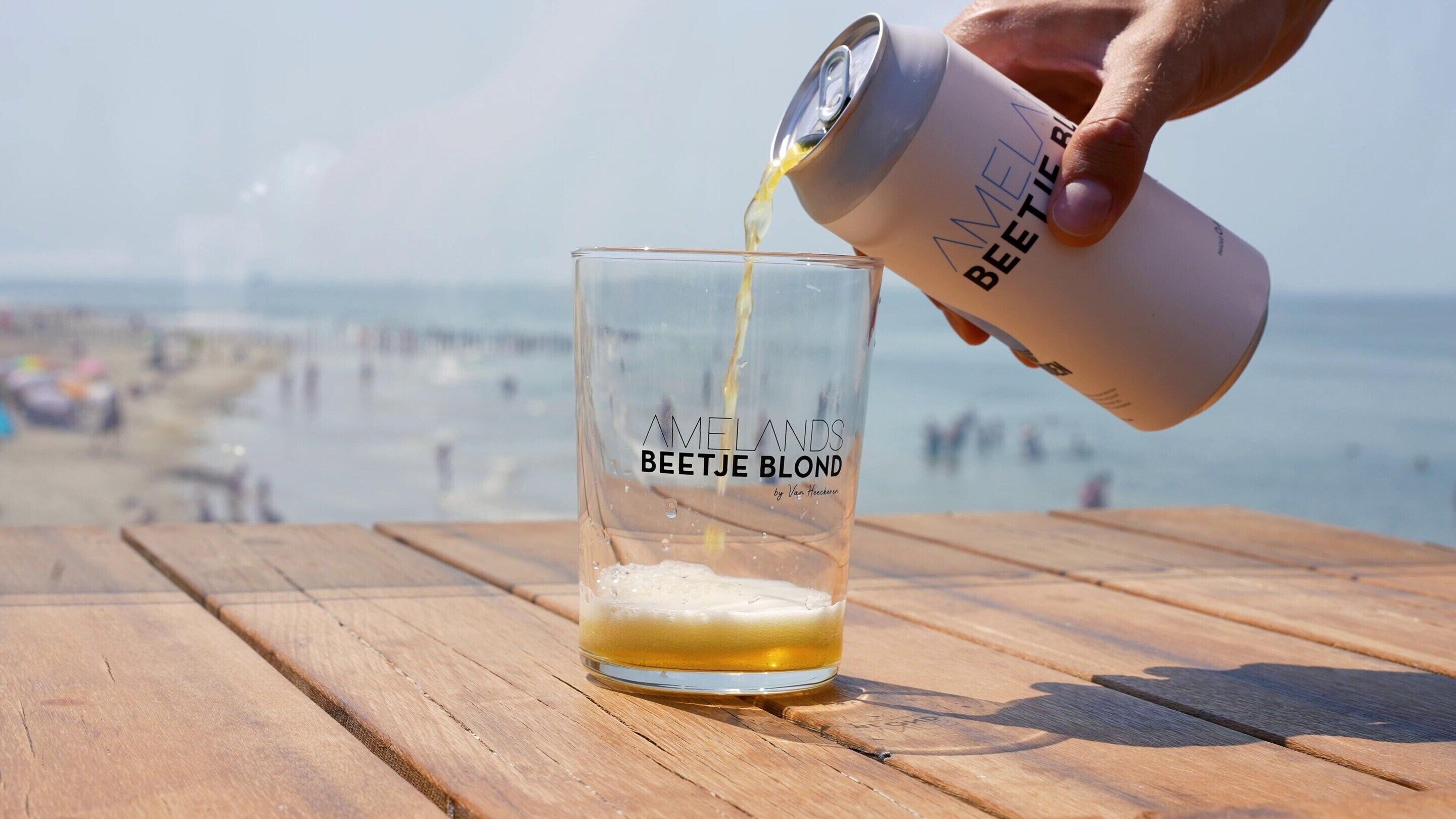 A glass labeled "Amelands Beetje Blond" being filled with a beverage on a wooden table at a beach.
