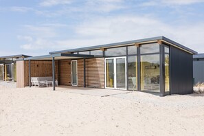 A modern building with a covered patio, large windows, and a sandy ground. - Comfy Lodge with Microwave Oven near Beach (Hollum)