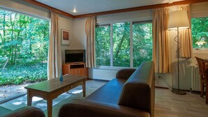 A living room with a sofa, TV, and a table. There are large windows with curtains, a radiator, and a lamp. - Chalet Near Trails & Town (Epe)