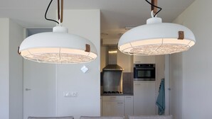 Uma área de refeições com mesa de madeira e quatro cadeiras cinzas, duas luminárias pendentes acima e vista para uma cozinha com eletrodomésticos modernos.