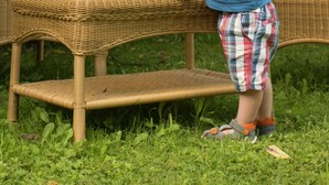 A child standing by a wicker table with a bottle and bowls, outdoors with greenery in the background.