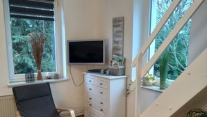 A room with a television, a white dresser, a chair, and a window with a view of trees.