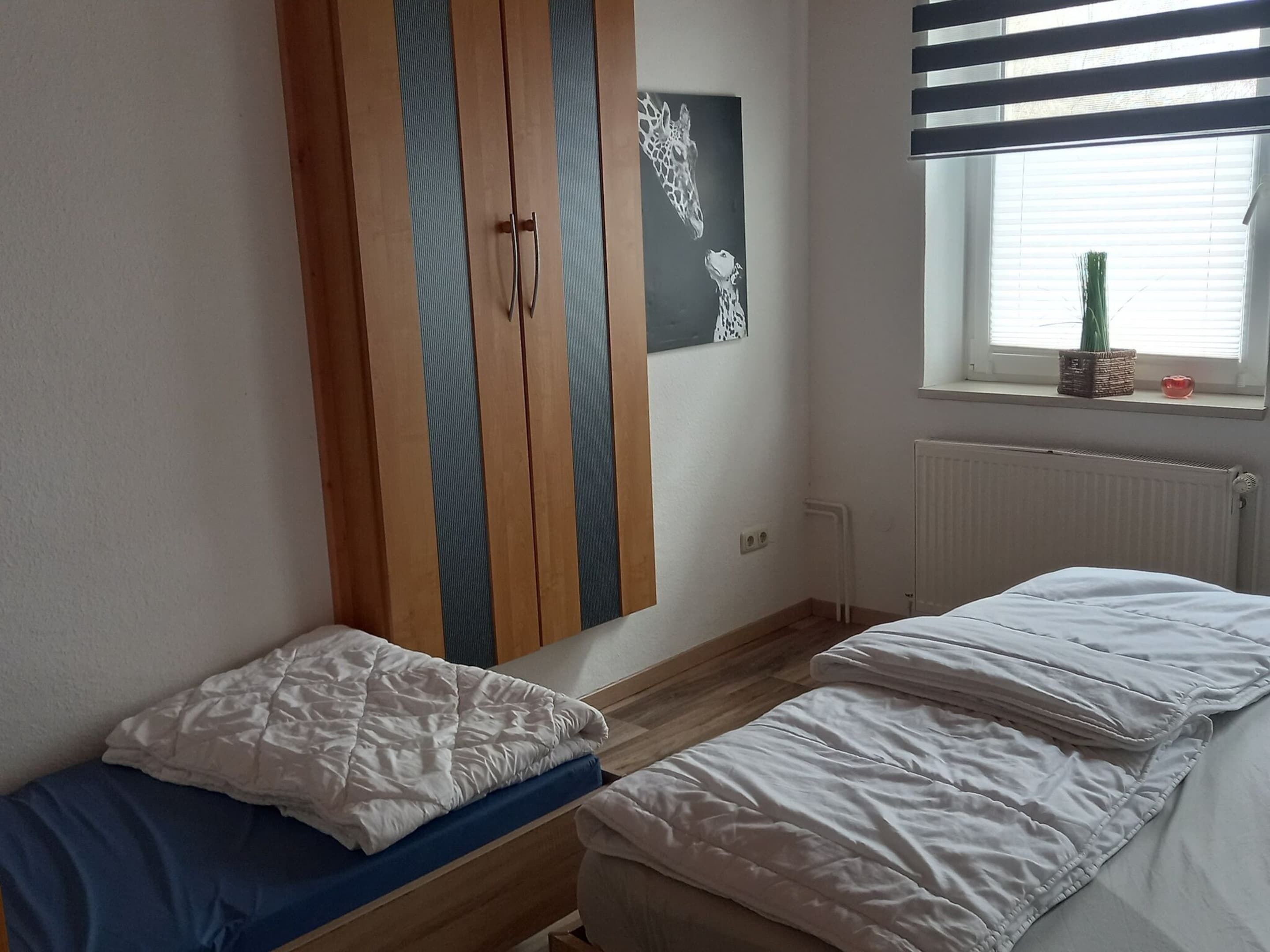 A bedroom with two single beds, a wardrobe with striped doors, a radiator, and a window with blinds.