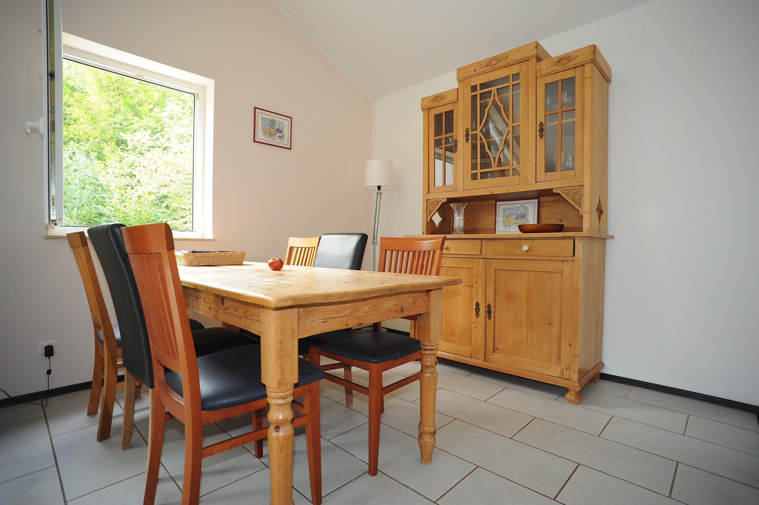 A dining area with a wooden table and chairs, a cabinet with glass doors, and a window with a view of greenery.