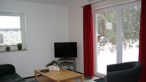Un salon avec une table basse en bois, un canapé noir, un fauteuil noir, un téléviseur et une fenêtre avec des rideaux rouges.
