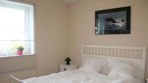 Une chambre avec un lit blanc, une table de chevet avec une lampe, une fenêtre avec des stores et un tableau encadré au mur.