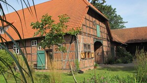 A traditional half-timbered house with a red-tiled roof, a wooden door, and a small garden with grass and flowers.