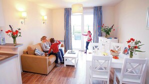 A living room with a sofa, a dining table with chairs, a kitchen area, and a view through a window. - Spacious Coastal Apt w/ Garden (Le Crotoy)