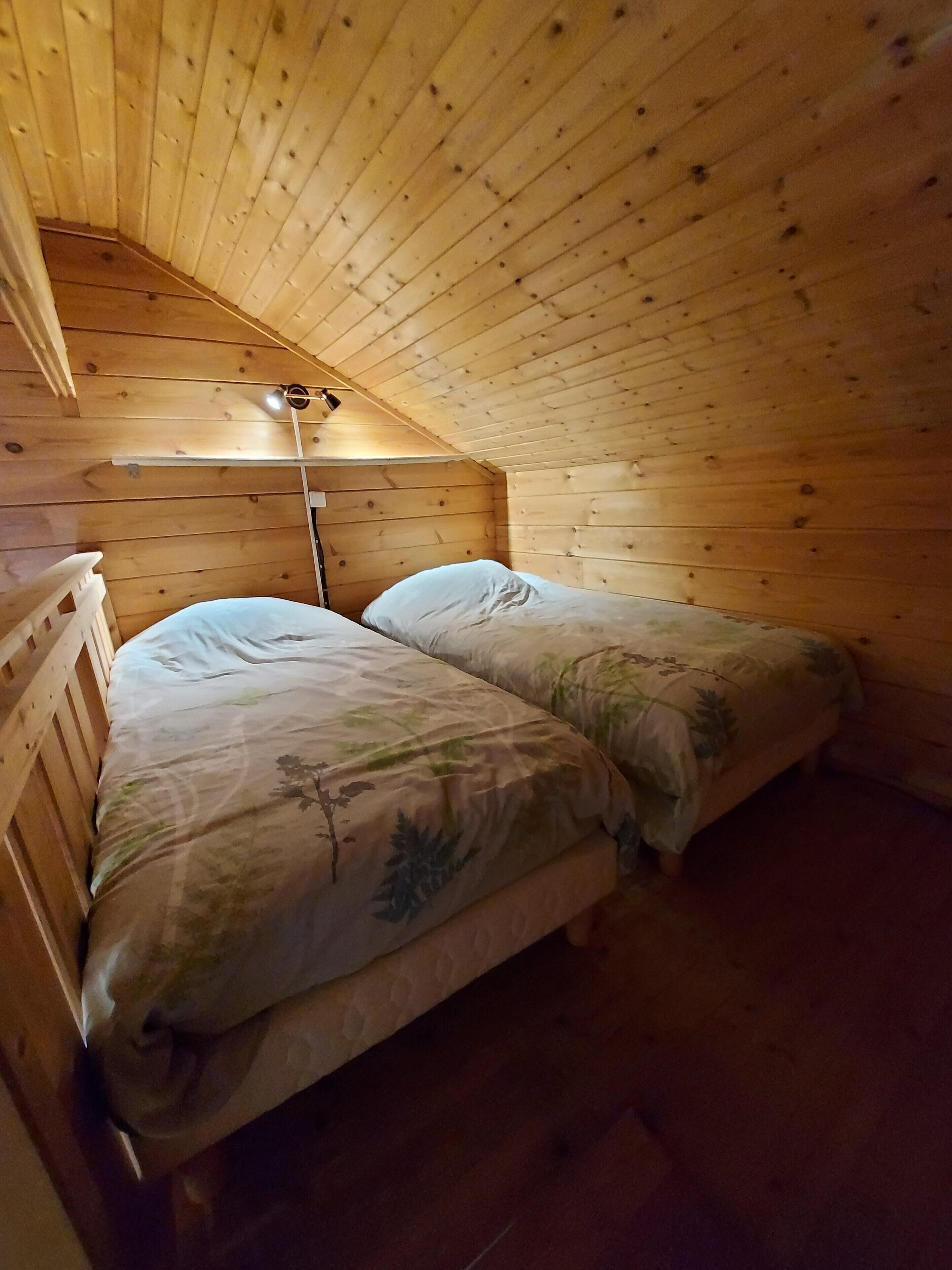 A small, cozy room with wooden walls and a sloped ceiling, featuring two beds with floral bedding and a single bedside table.