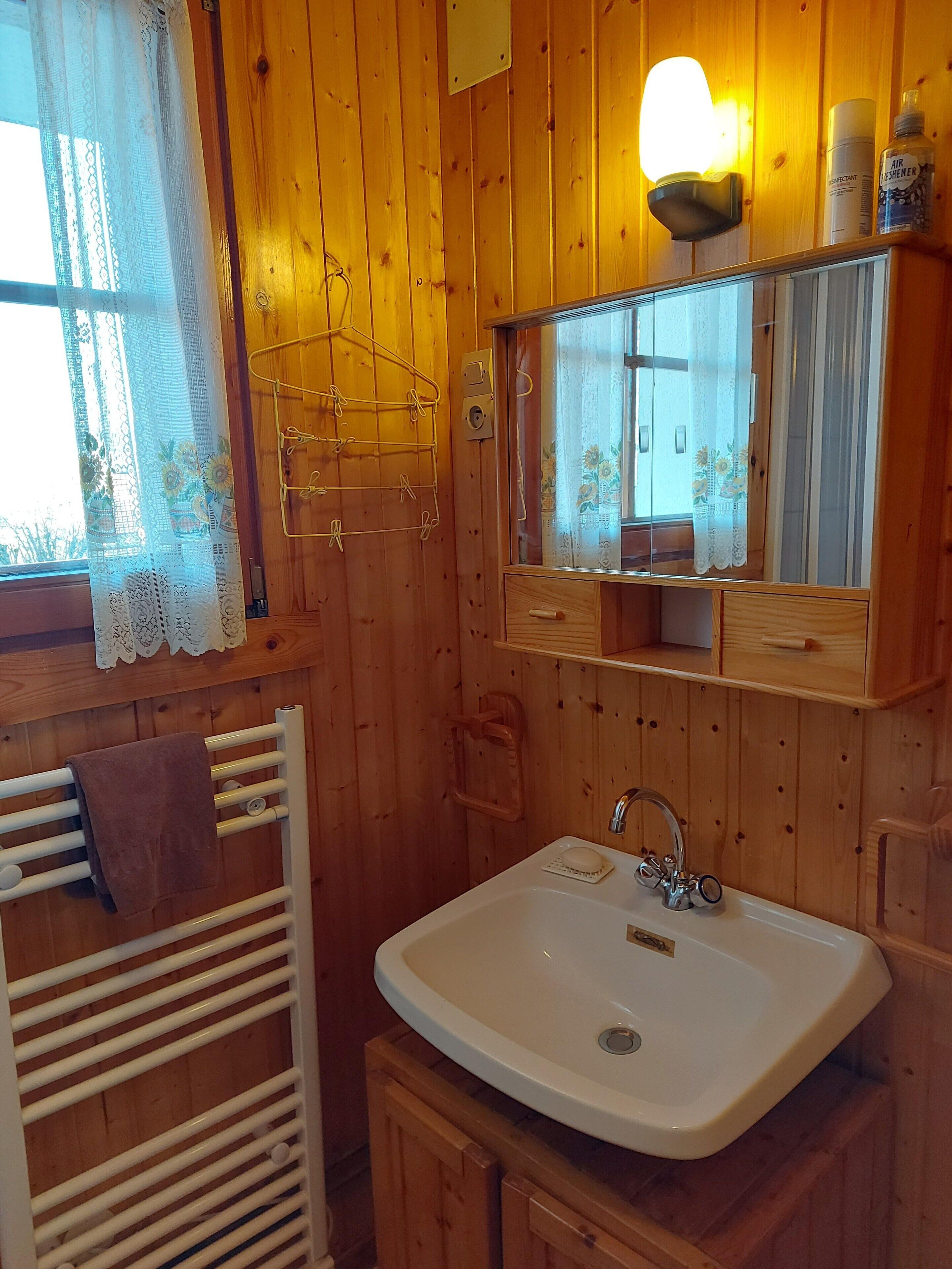 Ett badrum i en stuga med träpanel, ett vitt handfat, en handduksställning, en spegel och ett fönster med spetsgardiner.