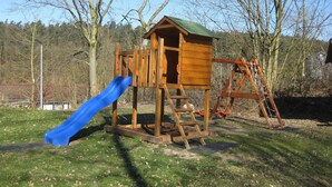 A wooden playhouse with a slide and climbing ladder in a grassy park area. - Holiday Home in Germany With Idyllic Nature (Ronshausen)