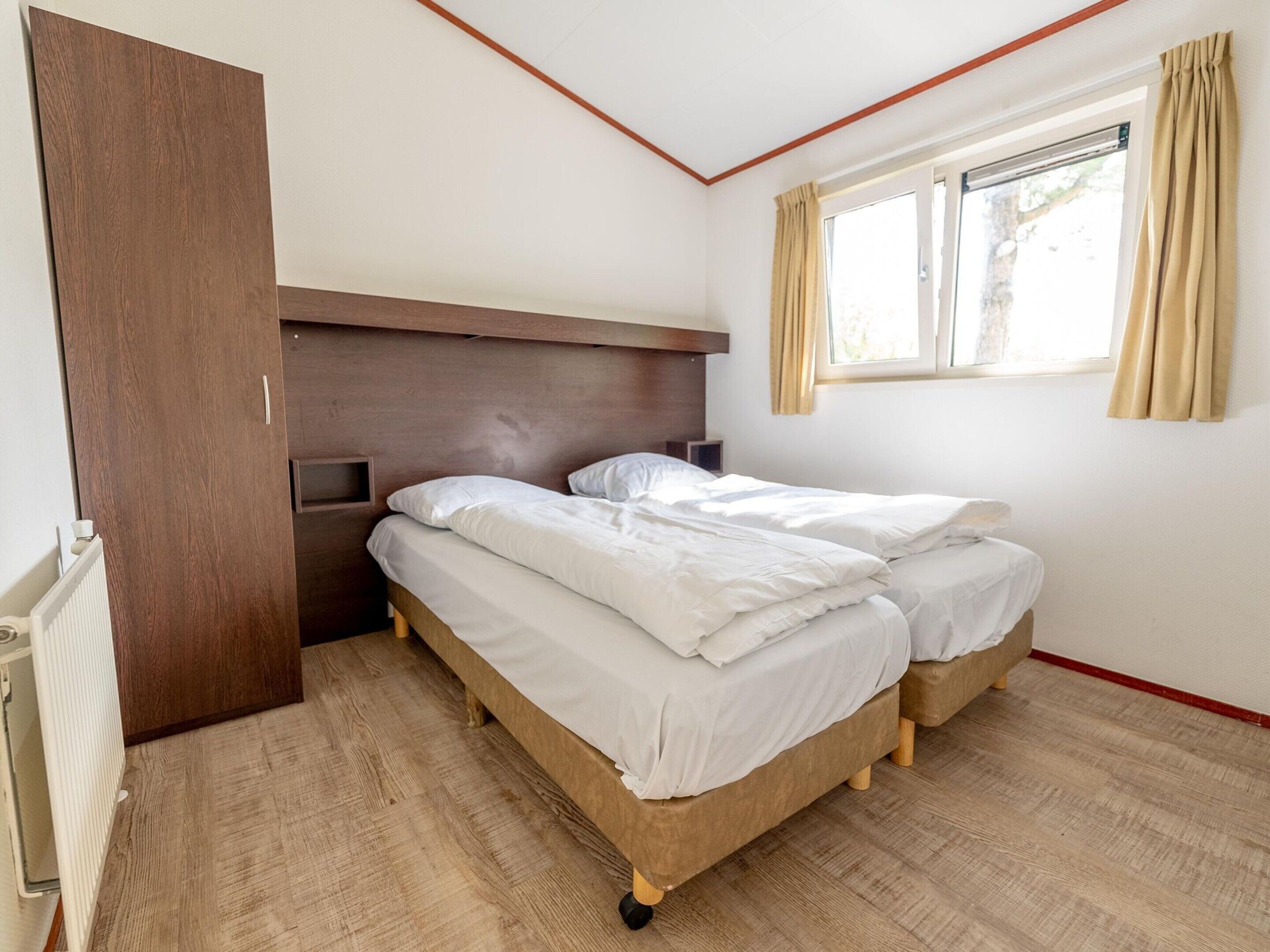 A bedroom with two beds, a wooden headboard, a window with curtains, and a radiator.