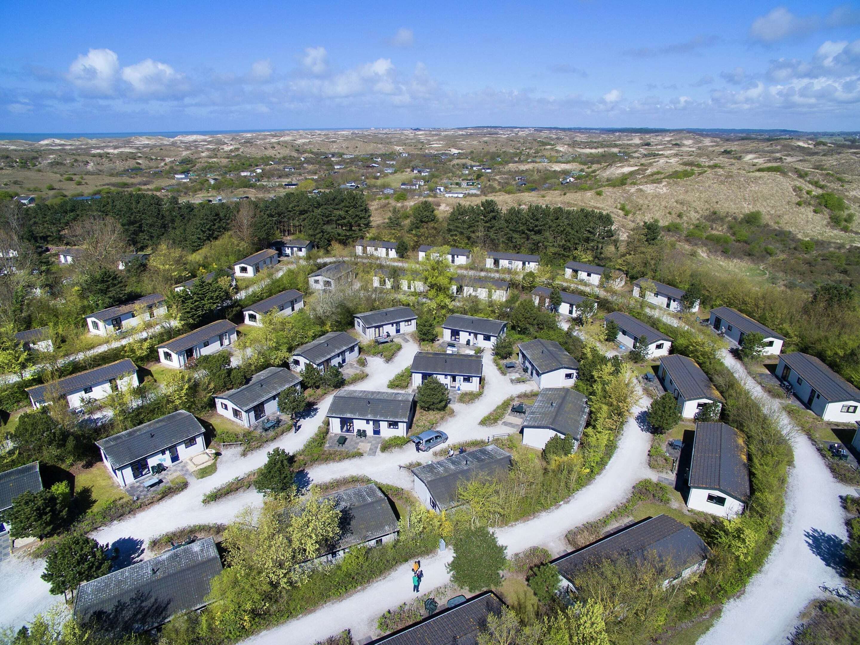 Distribución circular de pequeñas cabañas de una sola planta con un camino de grava, rodeado de vegetación y una vista distante del mar.