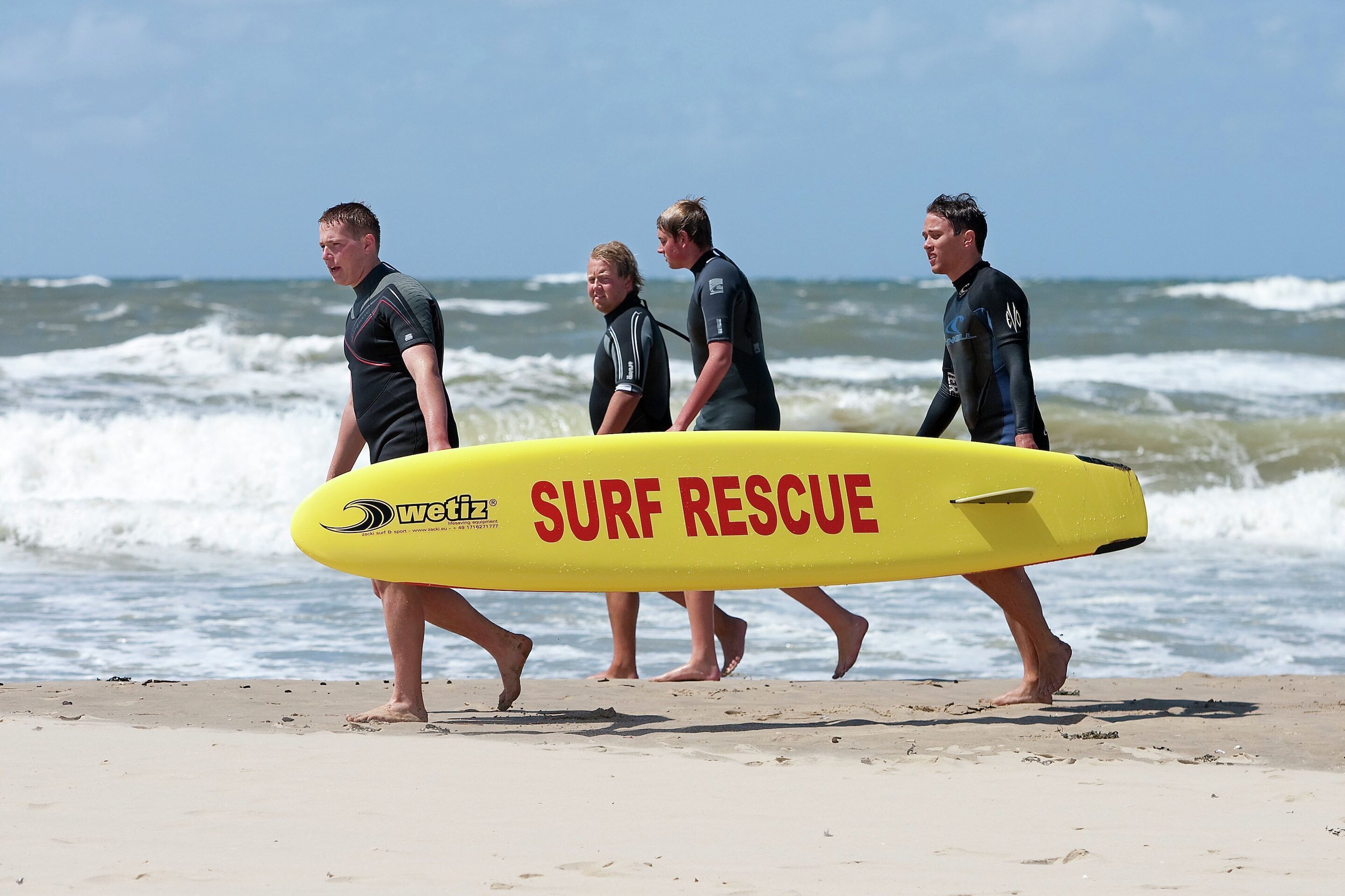 Four individuals walking on a beach, each carrying a yellow surfboard with 'SURF RESCUE' written on it.