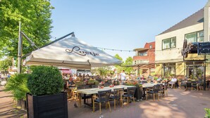 An outdoor café with tables and chairs under a large umbrella, surrounded by brick buildings and greenery. - Family Chalet in Netherlands Holiday Park (Egmond aan den Hoef)