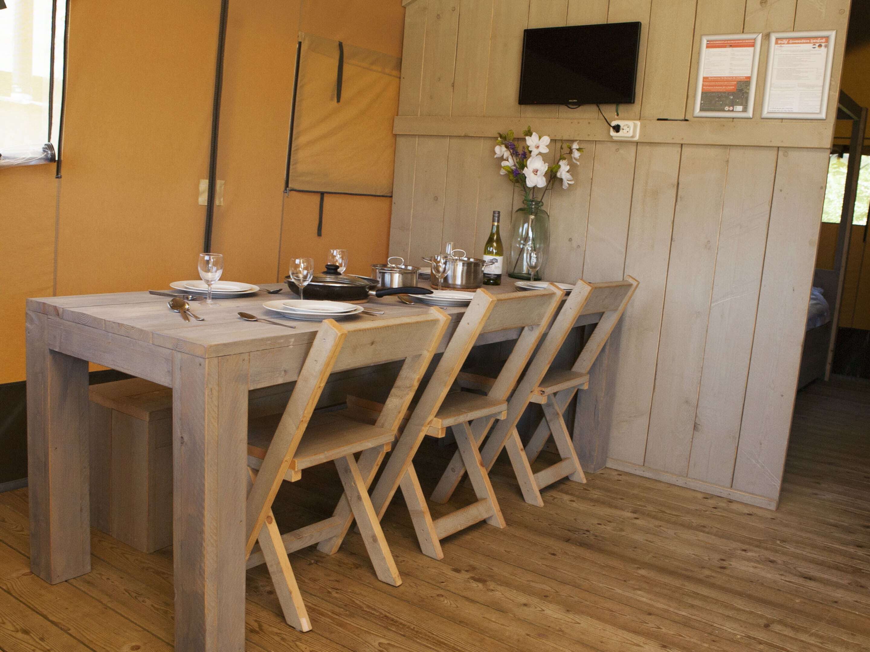 A wooden dining table with four chairs, a TV mounted on the wall, and a small vase with flowers.