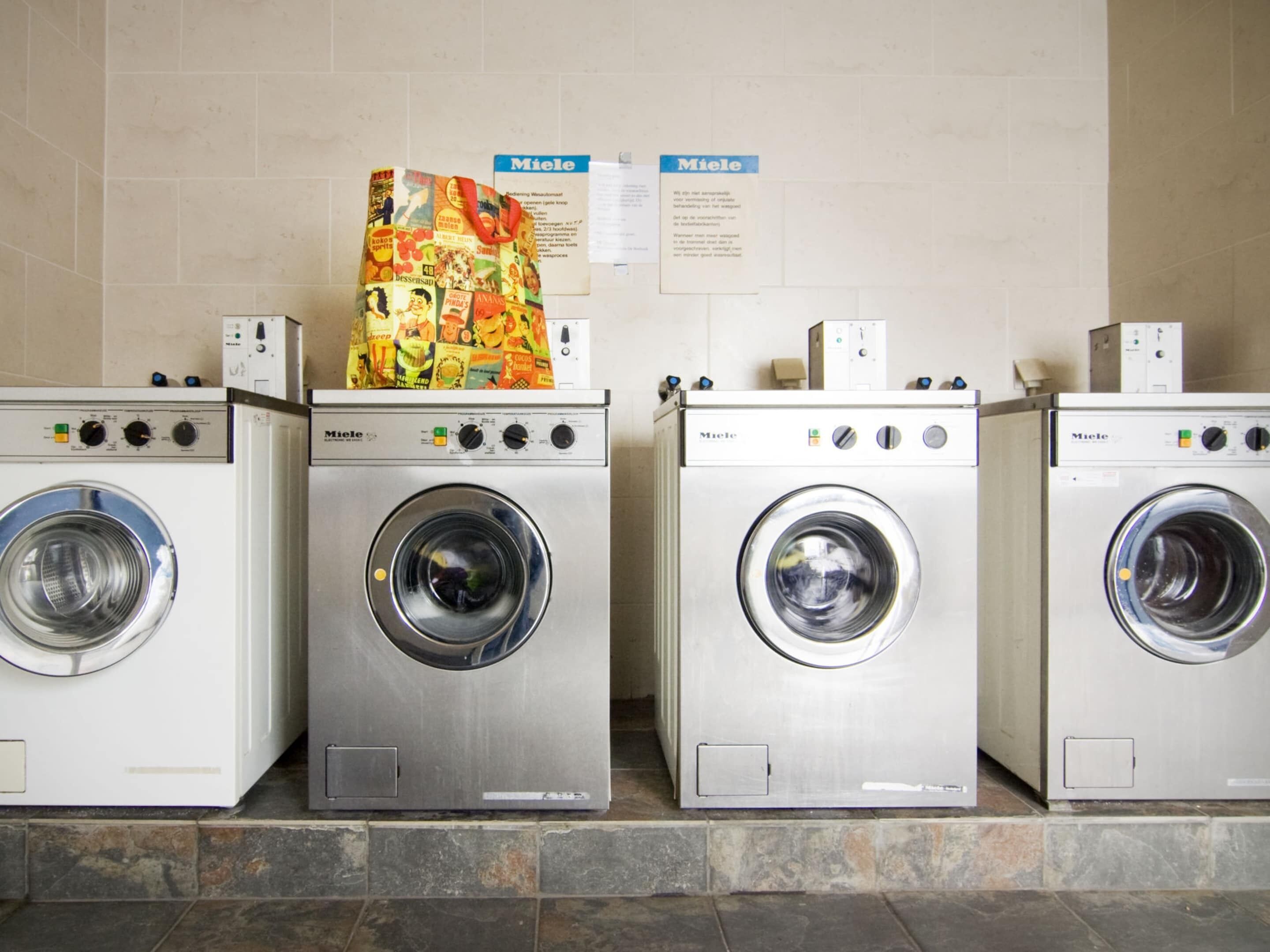 A row of identical front-loading washing machines with control panels and a tiled wall in the background.
