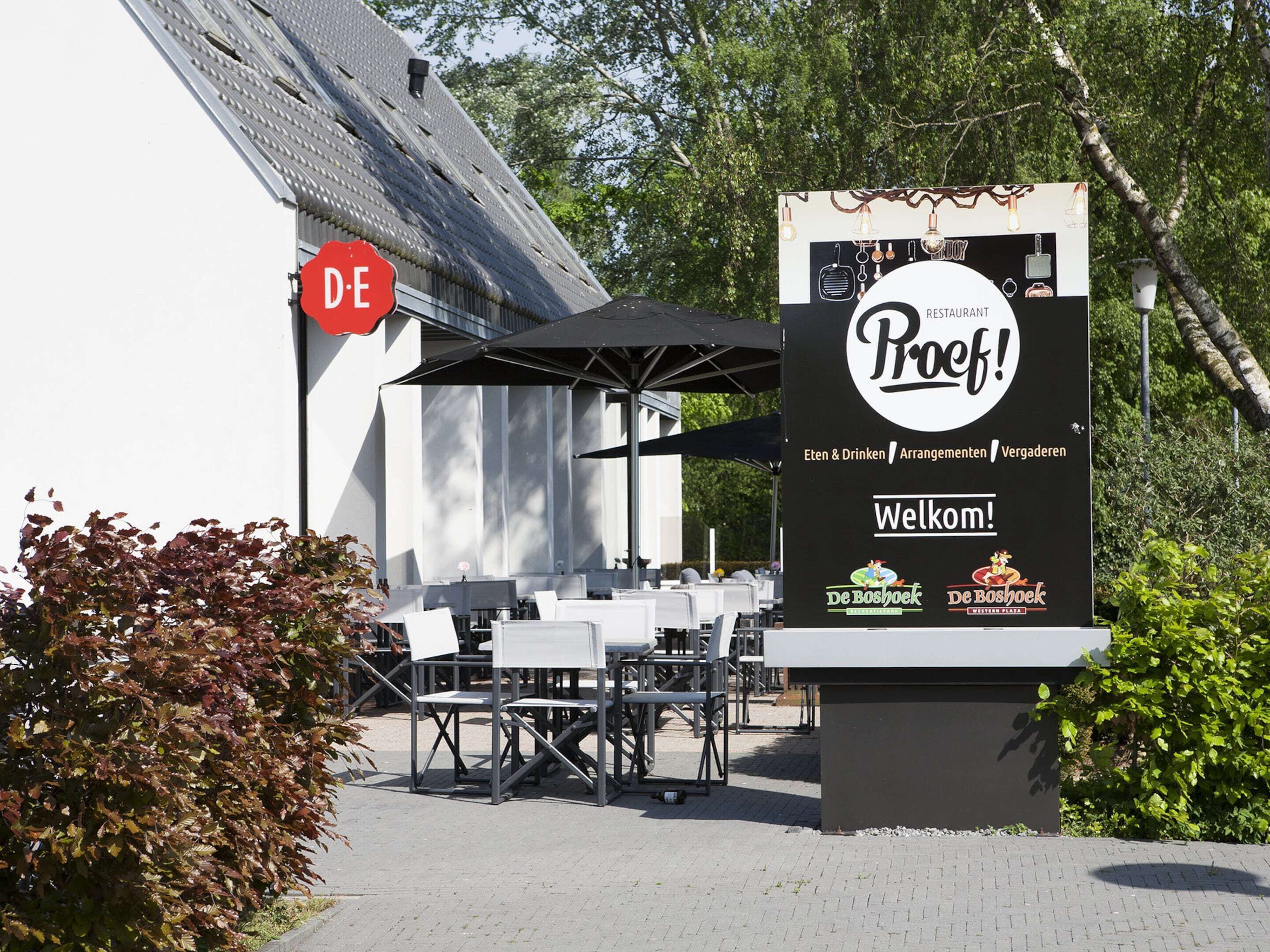 An outdoor seating area with tables and chairs, a sign for 'De Boshoek Restaurant', and a red 'DE' sign.