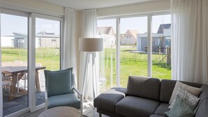 A modern living room with a grey sofa, a blue armchair, and a round wooden coffee table. Large windows offer a view of a garden and neighboring houses. - Bungalow at Nieuwvliet Bad Beach Resort (Nieuwvliet)
