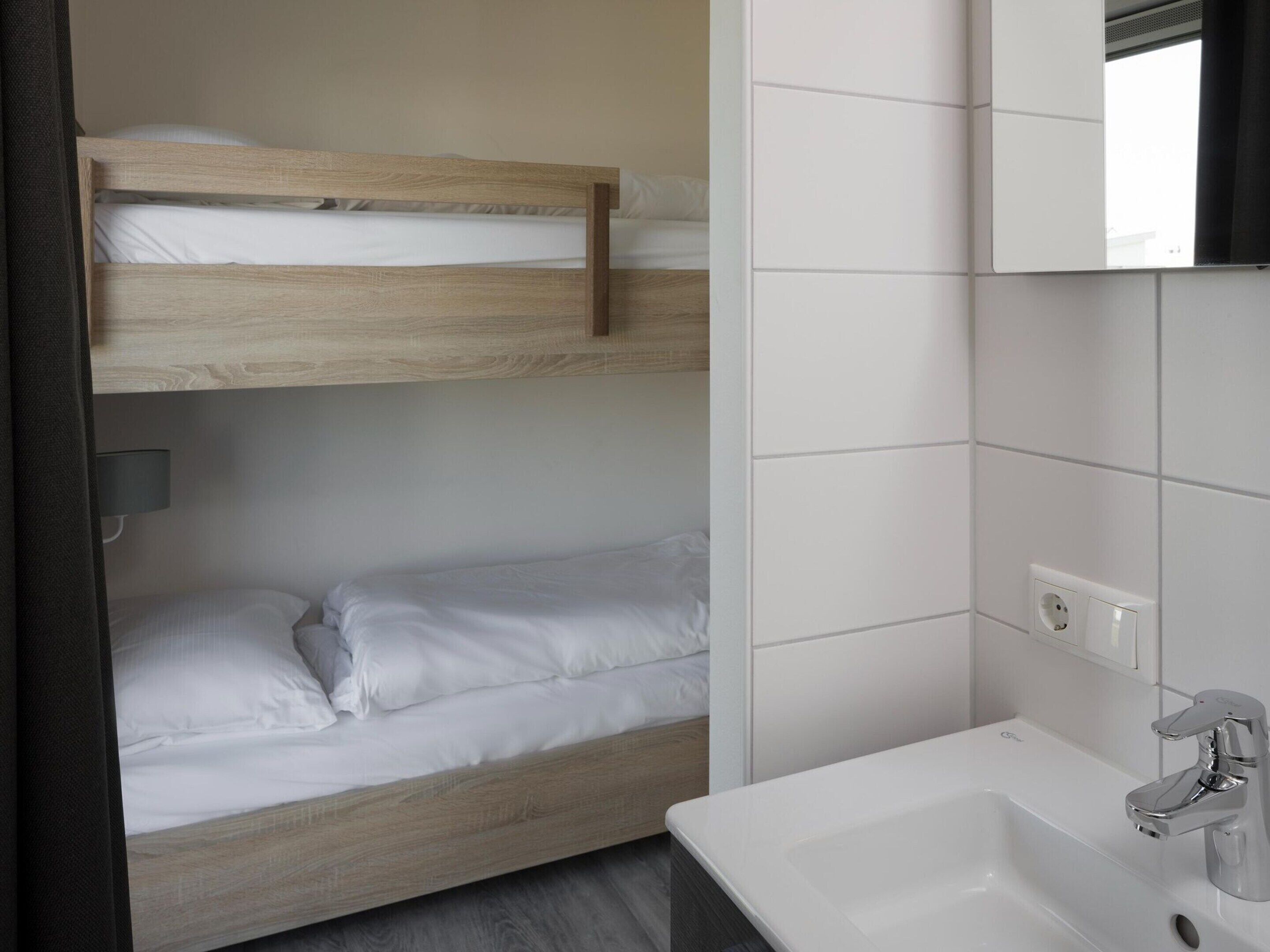 Ein Stockbett mit weißer Bettwäsche, ein weißes Waschbecken mit chromfarbenem Wasserhahn und ein Spiegel über dem Waschbecken.