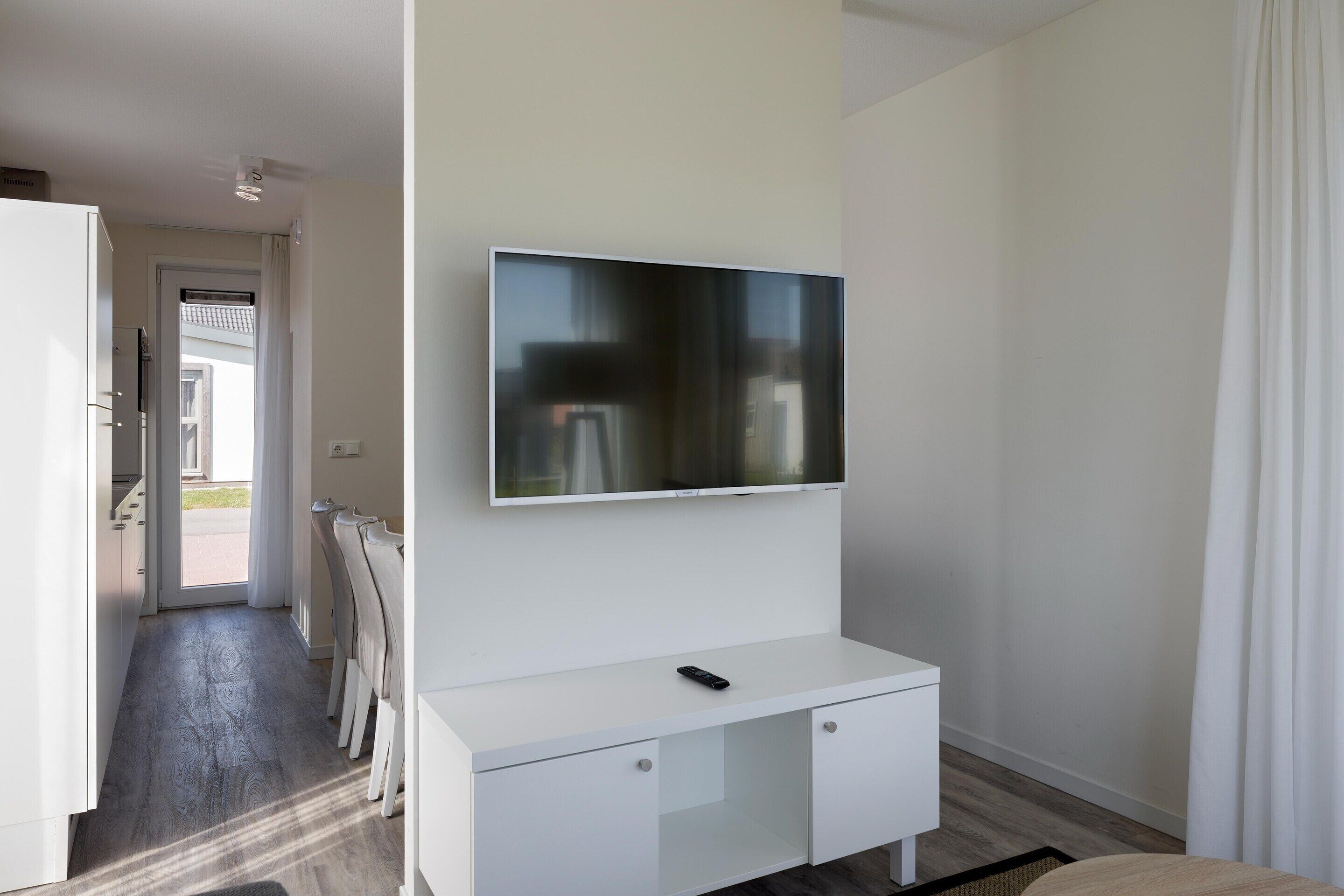A modern room with a flat-screen TV mounted on a white wall, a white console table with a remote, and a view of the outside through a door.