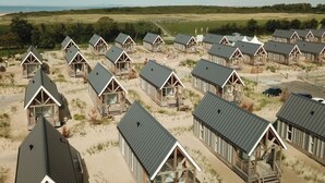 Eine Reihe von Holzstrandhäusern mit Metalldächern, angeordnet auf sandigem Gelände mit Blick auf das Meer und die entfernten Hügel.
