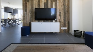 A modern living room with a large flat-screen TV mounted on a wooden panel wall, a white entertainment unit, and a dining area with a glass table and chairs. - Villa in Cadzand-bad Near North Sea Coast (Cadzand-Bad)