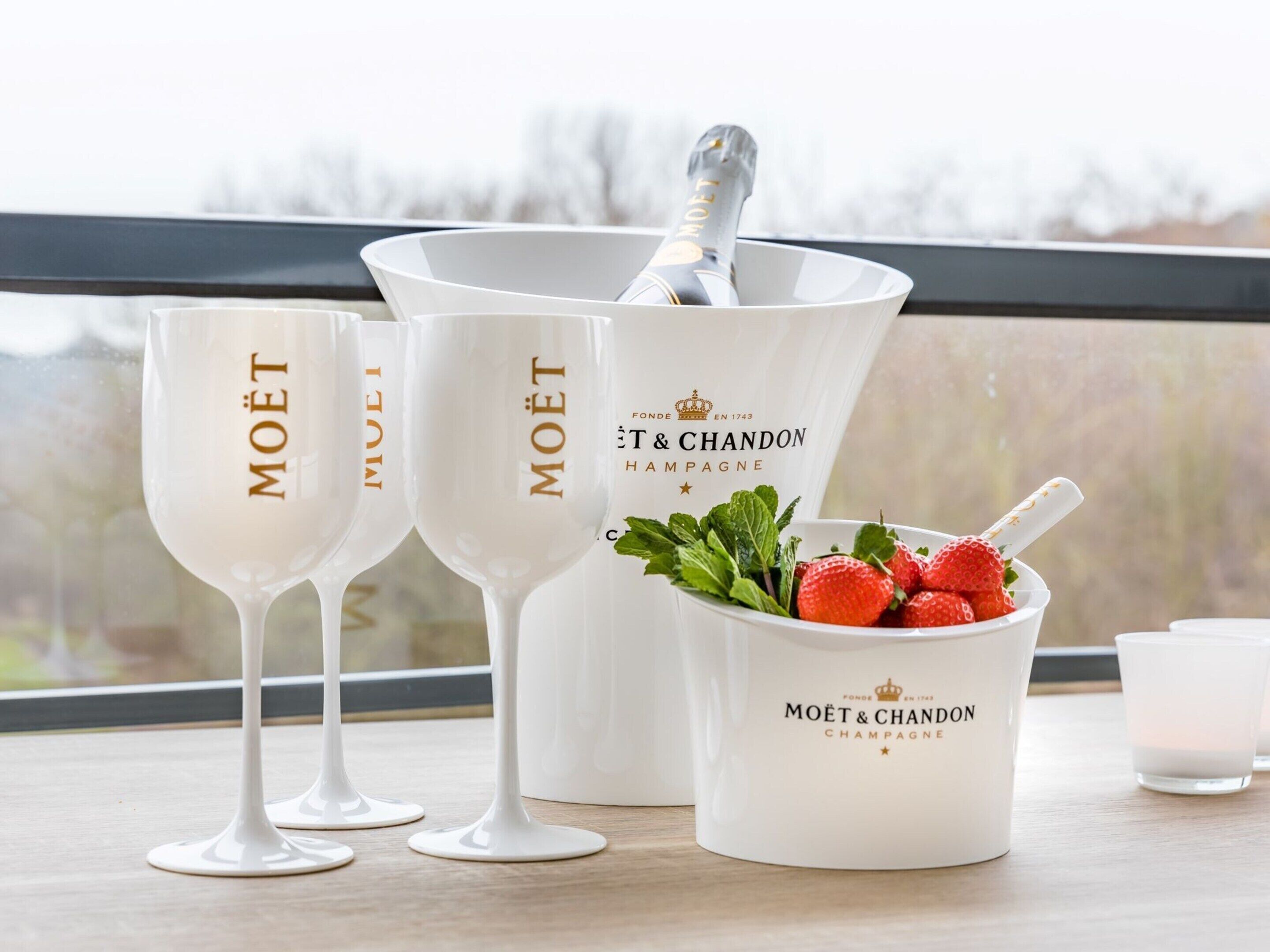 Three white wine glasses with "MOËT" branding, a white bucket with a champagne bottle and strawberries, and a white mug with "MOËT & CHANDON CHAMPAGNE" branding.