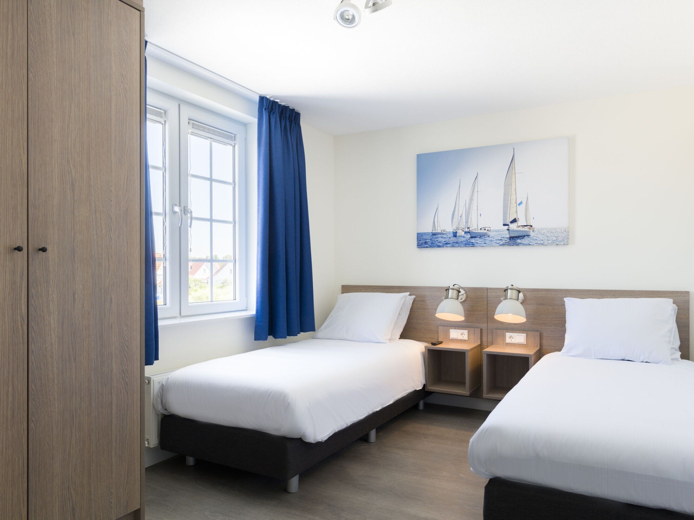 Habitación de hotel con dos camas, una ventana con cortinas azules, un cuadro de veleros y lámparas de noche.