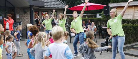 An outdoor gathering with people in green shirts, some children, and a building with a red umbrella in the background.
