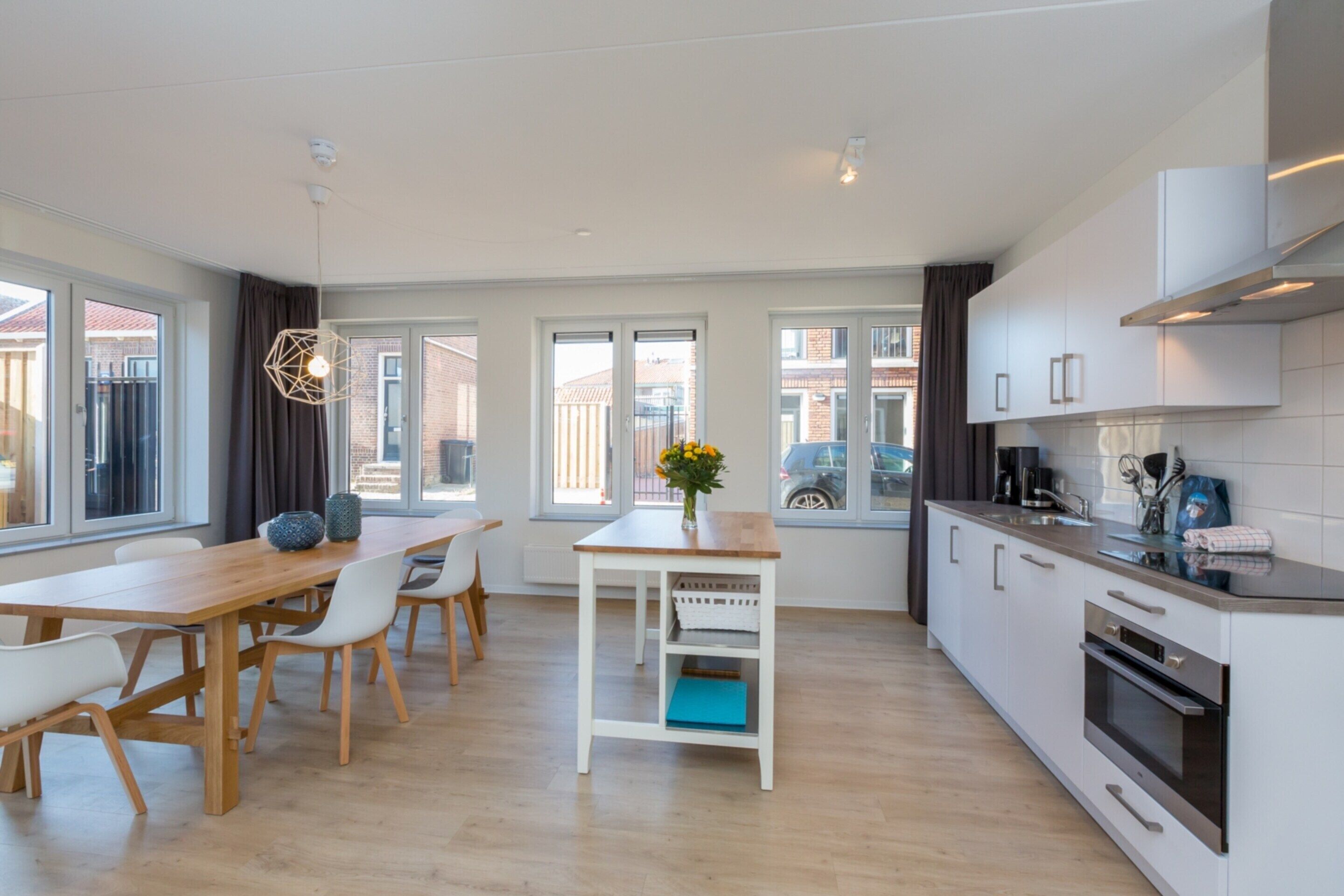 A modern kitchen with white cabinets, a wooden dining table, and a view of the street.