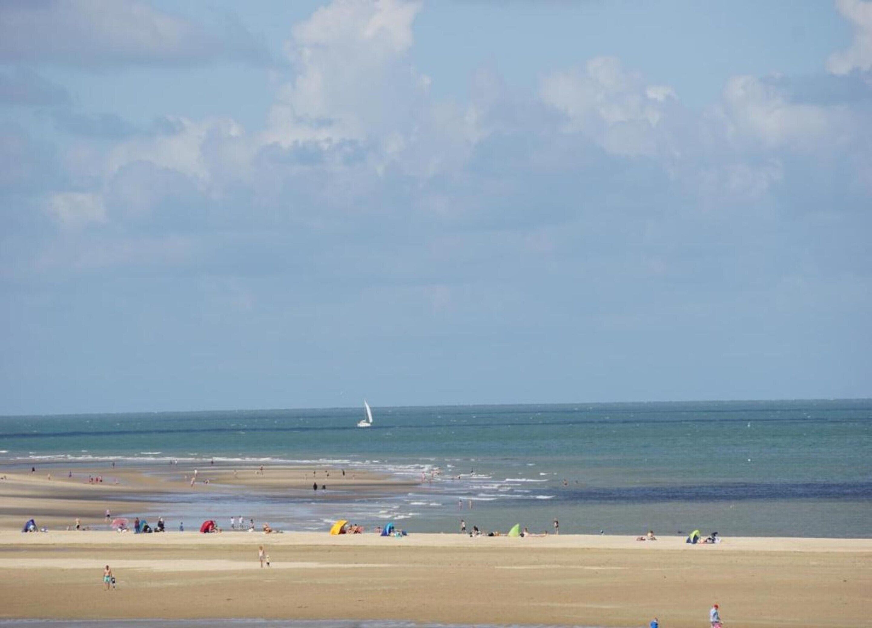 Une plage avec un voilier à l’horizon, des gens sur le sable et des tentes.