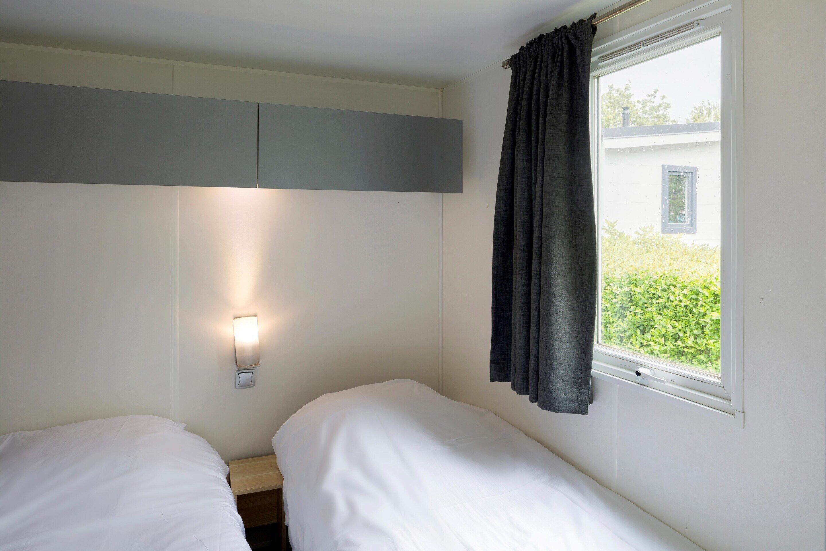 A single-bed room with a bedside table, a wall-mounted light, and a window with curtains.