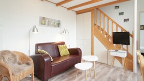 A furnished living room with a brown leather sofa, a wicker chair, a white table, and a television on a stand. - Cozy Apartment near Sea in Bergen aan Zee (Bergen aan Zee)