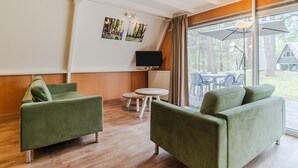 Ein Zimmer mit zwei grünen Sofas, einem kleinen Tisch, einem Fernseher und einem großen Fenster mit Blick auf Bäume.