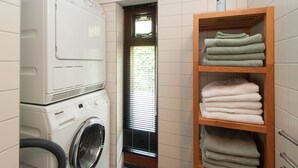 Ein Waschraum mit nebeneinander gestapelten Waschmaschine und Trockner, einem Fenster mit Jalousien und einem Holzregal mit gefalteten Handtüchern.