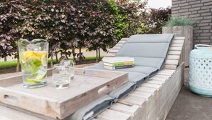 Eine Holzterrasse mit Liegestuhl, einem Tablett mit Getränken und Büchern sowie einem Blumentopf.