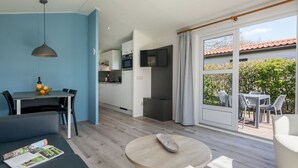 Un salon moderne avec un canapé, une table basse ronde et un coin repas comprenant une table et des chaises. Au fond, on aperçoit une cuisine avec des meubles blancs et un téléviseur fixé au-dessus du comptoir.