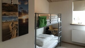 Ein Schlafraum mit Etagenbetten, Holzboden und Heizkörper. An der Wand hängen zwei Bilder, eines zeigt einen Leuchtturm, das andere einen Sonnenuntergang.
