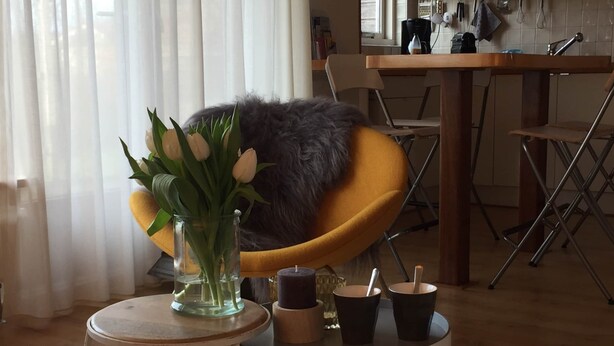 Ein gemütliches Wohnzimmer mit einem gelben Sessel, einem kleinen runden Tisch mit Blumen und einem Küchenbereich im Hintergrund.