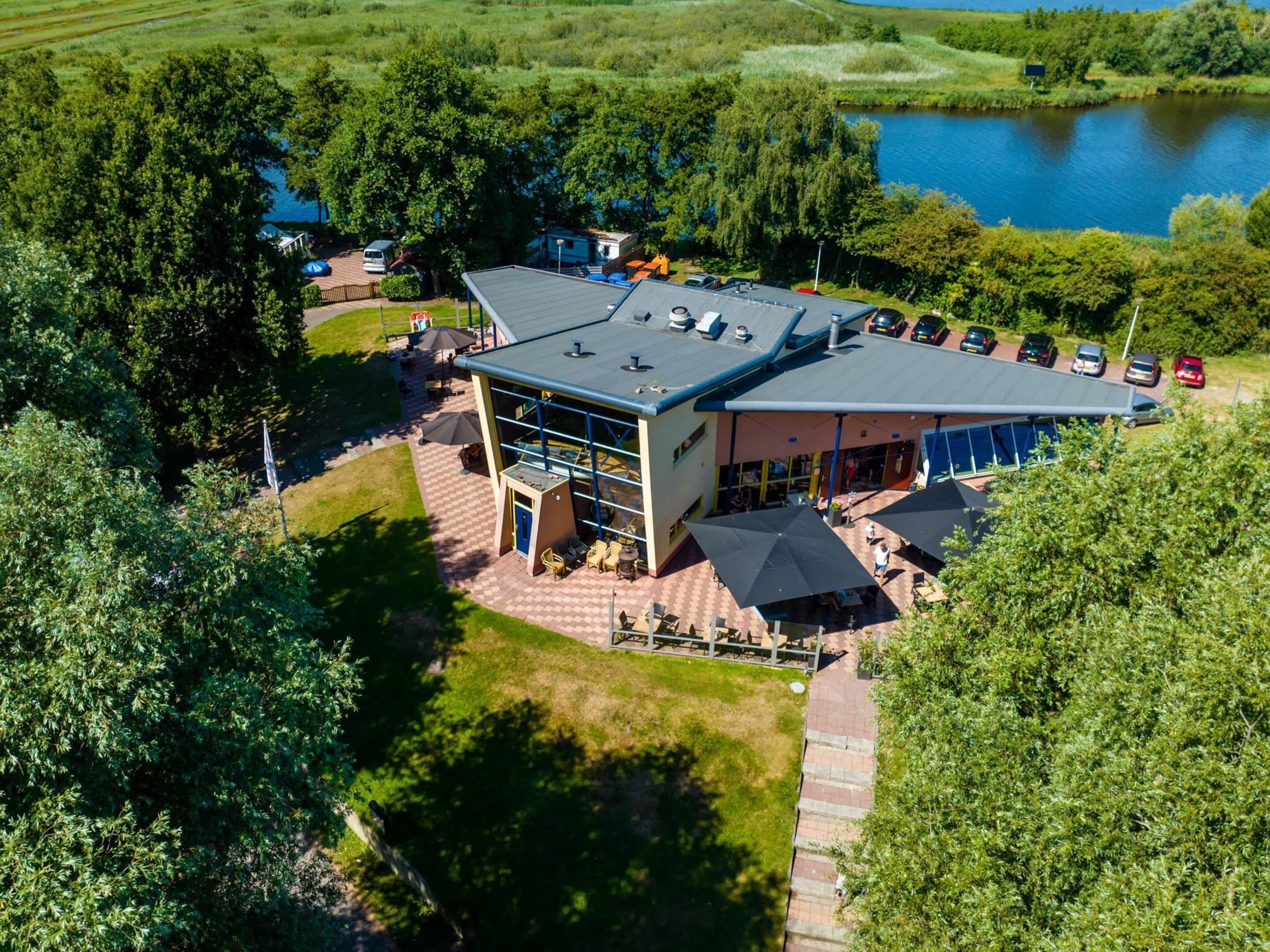 An aerial view of a modern building with a flat roof, surrounded by lush greenery and a body of water.