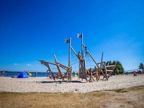 A beach with a large wooden pirate ship structure, a blue tent, and people engaging in activities.