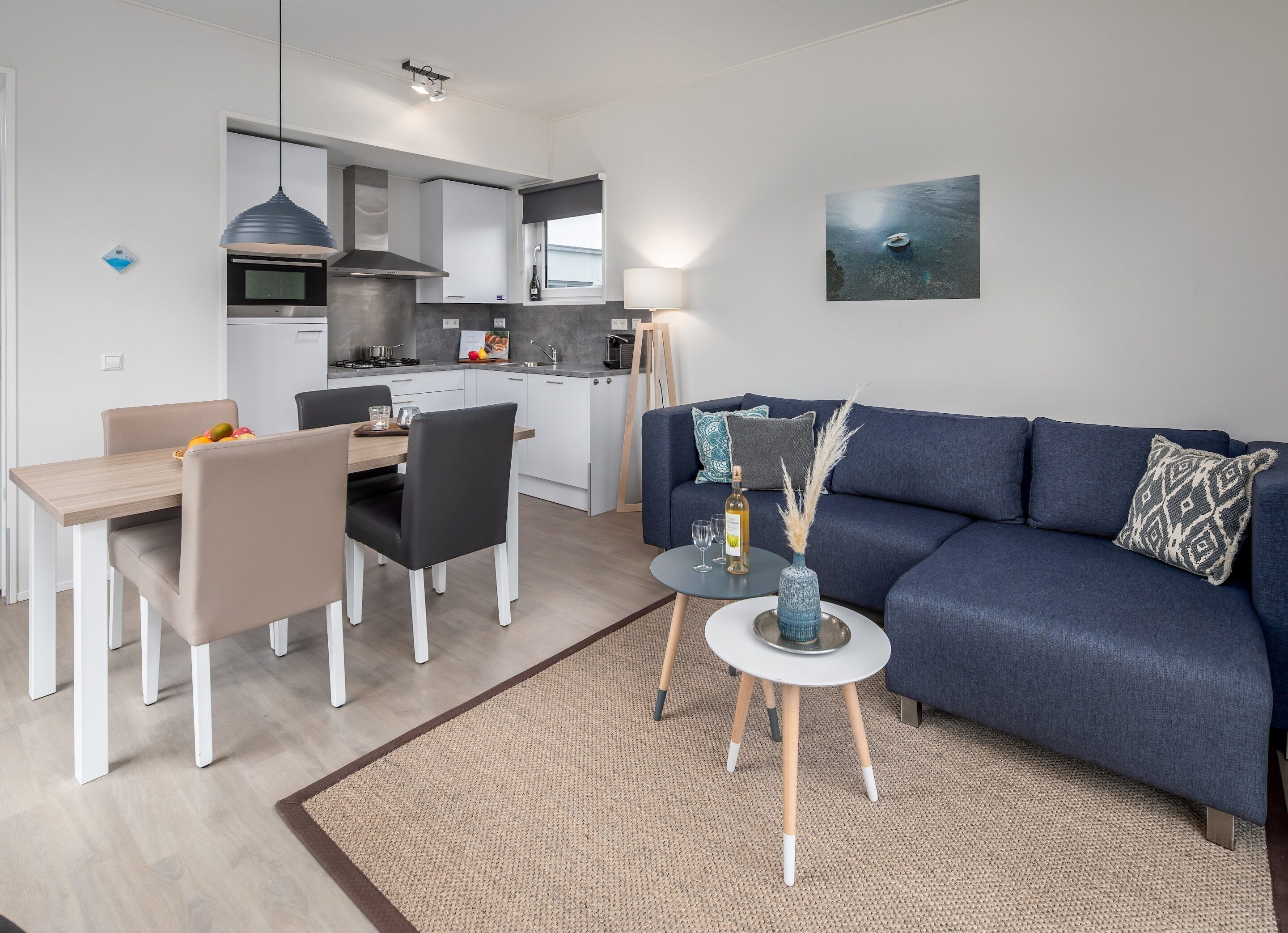A modern living room with a blue sofa, a dining table with chairs, a kitchen area, and a small side table with a vase.