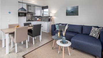 A modern living room with a blue sofa, a dining table with chairs, a kitchen area, and a small side table with a vase.