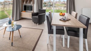 A modern living room with a dining table, grey armchairs, a TV, and a view of a residential area. - Cozy Chalet Near Breskens Sea (Breskens)