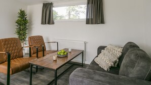 Una sala de estar con un sofá, dos sillones, una mesa de centro con un cuenco de manzanas, una planta en maceta y una ventana con cortinas.