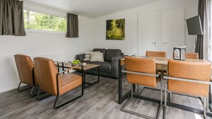Una sala de estar moderna con sofá, mesa de centro y mesa de comedor con sillas. Hay un cuadro en la pared, un televisor montado y una ventana con cortinas.